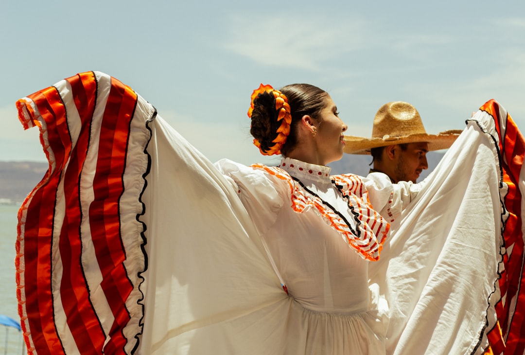 A female folkloric ballet dancer holding up her white and orange traditional dress alongside a male dancer wearing a sombrero under a clear blue sky at the boardwalk of lake Chapala in Jalisco, Mexico. by Hispanic Month A man and a woman dressed in mexican clothing